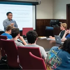 A group of students sitting around a boardroom style room during a presentation.